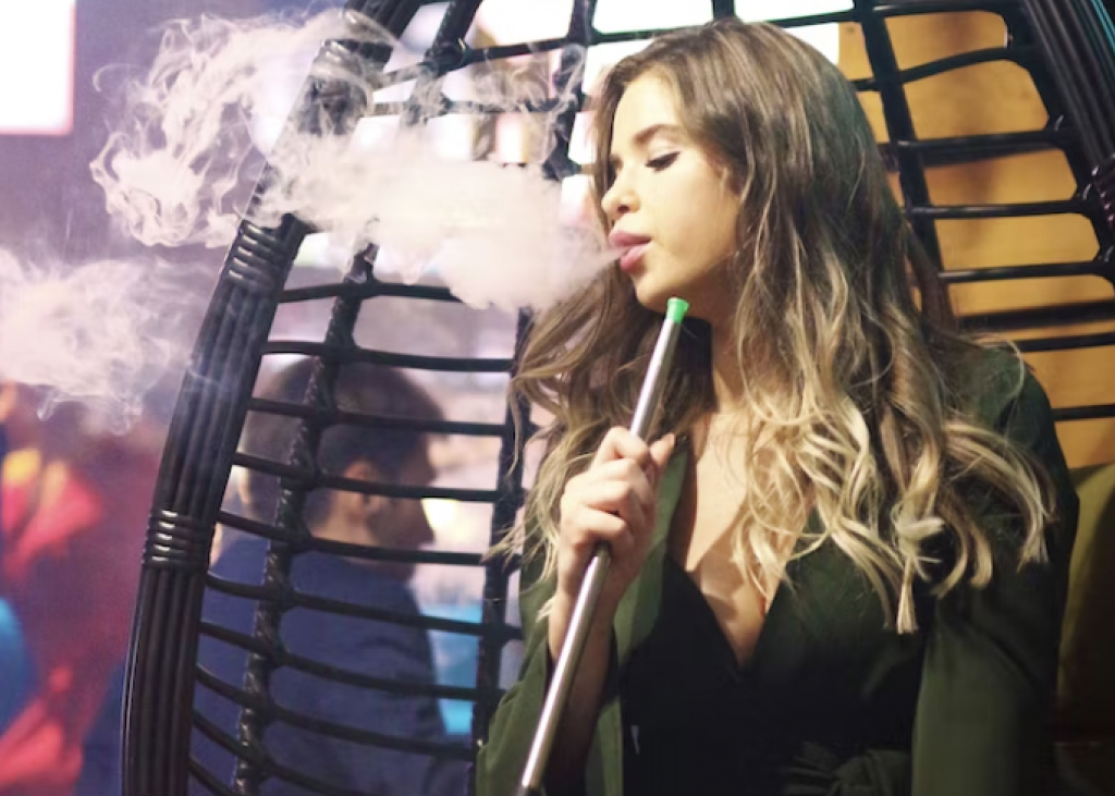 A person puffing out smoke with a blurry hookah lounge in the background.